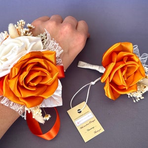 May include: A wrist corsage and boutonniere set with orange and white satin roses, white lace, and dried baby's breath. The tag reads "With love from [Name] Charming - Thank you!"