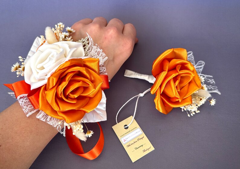 May include: A wrist corsage and boutonniere set with orange and white satin roses, white lace, and dried baby's breath. The tag reads "With love from [Name] Charming - Thank you!"