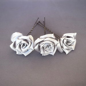 May include: Three silver satin rose hair clips with wire stems. The roses are arranged in a row, with the stems pointing towards the right.