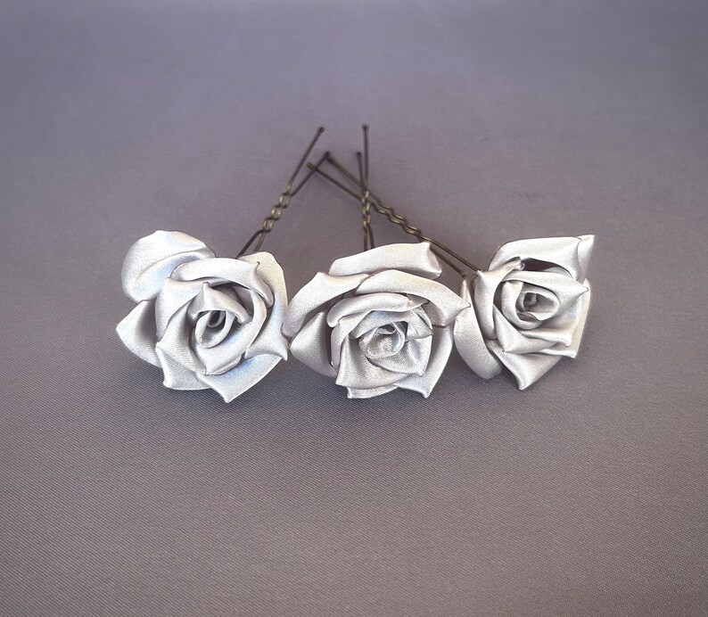 May include: Three silver satin rose hair clips with wire stems. The roses are arranged in a row, with the stems pointing towards the right.