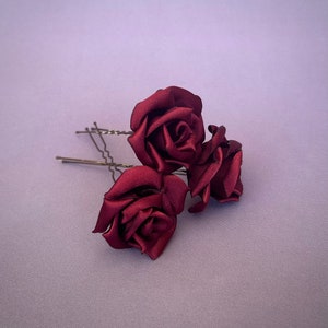 May include: Three burgundy satin roses attached to bobby pins. The roses are arranged in a cluster, with the largest rose in the center and the two smaller roses on either side.