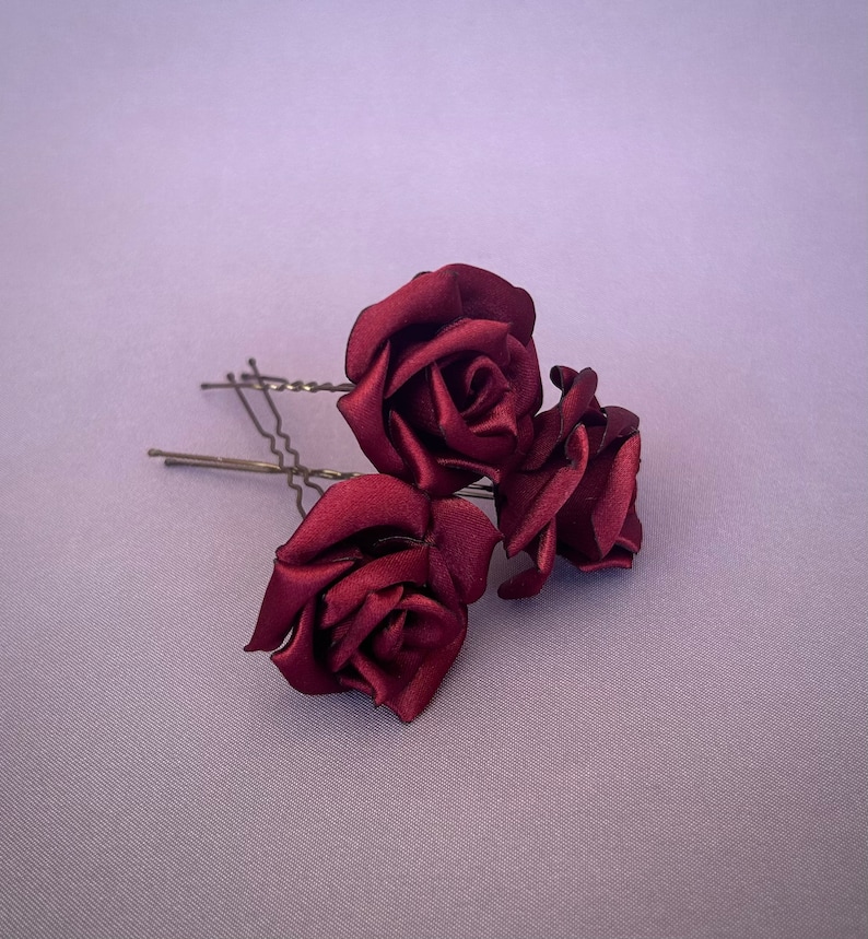 May include: Three burgundy satin roses attached to bobby pins. The roses are arranged in a cluster, with the largest rose in the center and the two smaller roses on either side.
