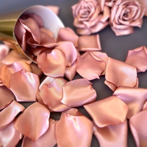 May include: A pile of rose petals made of satin fabric in a blush pink color. The petals are scattered on a gray surface. There are two rose flower decorations made of the same fabric in the background.