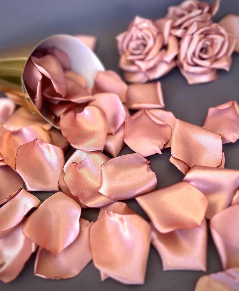 May include: A pile of rose petals made of satin fabric in a blush pink color. The petals are scattered on a gray surface. There are two rose flower decorations made of the same fabric in the background.