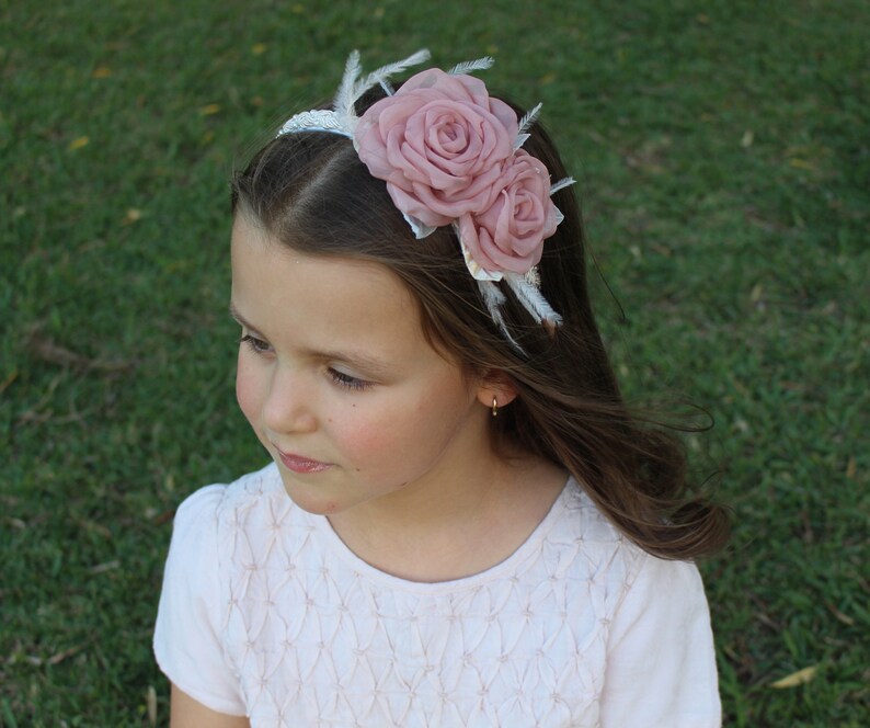 Op de afbeelding: Een wit hoofdband met twee roze stoffen rozen en witte veren. Het hoofdband wordt gedragen door een jong meisje met lang bruin haar.
