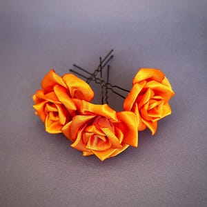 May include: Three orange satin roses attached to bobby pins. The roses are arranged in a cluster and are ready to be used as a hair accessory.