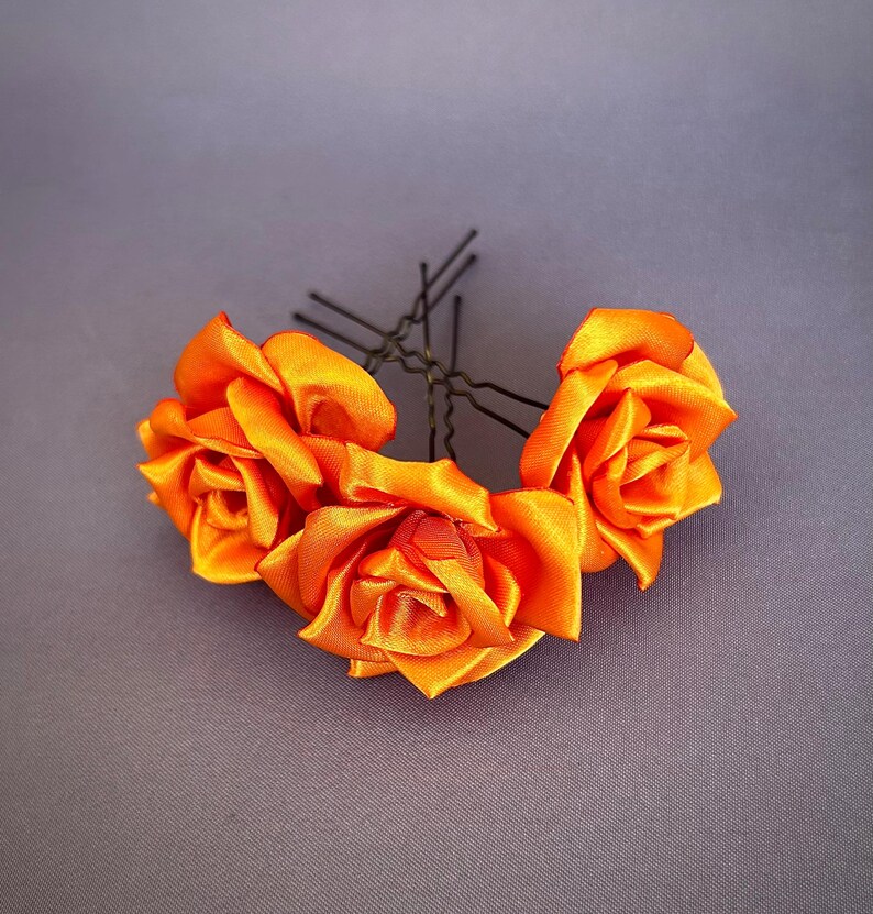 May include: Three orange satin roses attached to bobby pins. The roses are arranged in a cluster and are ready to be used as a hair accessory.