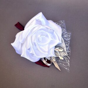 May include: A white satin rose boutonniere with a silver mesh and rhinestones.
