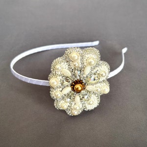 Puede incluir: Una diadema blanca con un gran adorno en forma de flor blanca. La flor est&aacute; hecha de cuentas blancas y perlas, con un centro dorado.