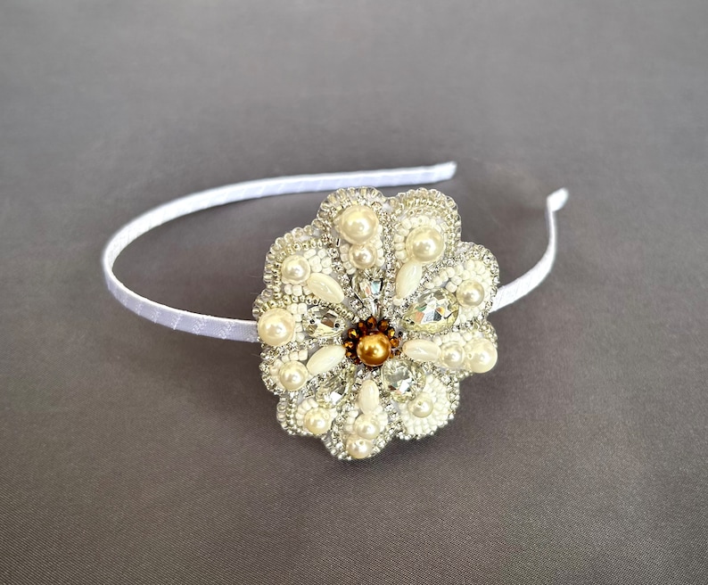 Puede incluir: Una diadema blanca con un gran adorno en forma de flor blanca. La flor est&aacute; hecha de cuentas blancas y perlas, con un centro dorado.