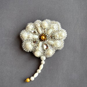Puede incluir: Un broche de flor blanca con cuentas de perla y plata con un centro dorado. El broche tiene un tallo largo hecho de perlas blancas.
