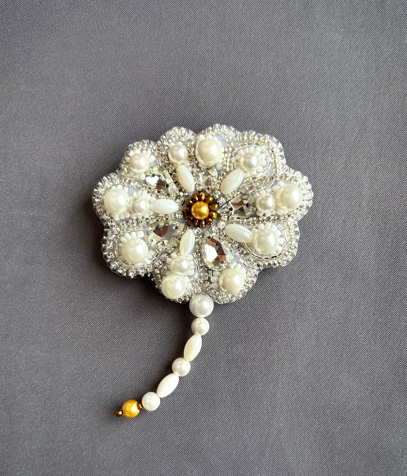Puede incluir: Un broche de flor blanca con cuentas de perla y plata con un centro dorado. El broche tiene un tallo largo hecho de perlas blancas.