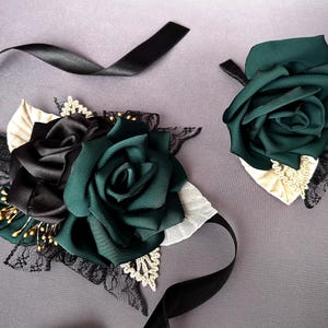 May include: Two satin rose hair accessories. One features a cluster of black and emerald green roses with lace and ribbon accents. The other is a single emerald green rose with cream and black lace. Both have black satin ribbons.