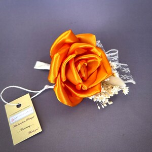 May include: A single orange satin rose with white lace and small white flowers. The rose is attached to a white ribbon. A small tag is attached to the ribbon with the text "Art Flowers" and "With love from Portugal... Charming... Thank you!"