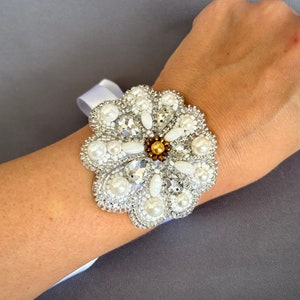 Puede incluir: Un brazalete de flores de cuentas blancas con detalles de perlas y un centro dorado. El brazalete se lleva en la mu&ntilde;eca.