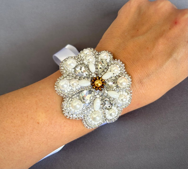 Puede incluir: Un brazalete de flores de cuentas blancas con detalles de perlas y un centro dorado. El brazalete se lleva en la mu&ntilde;eca.
