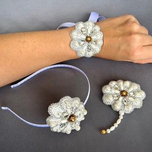 Puede incluir: Un conjunto de tres broches de flores blancas de perlas y oro. Un broche est&aacute; unido a una diadema blanca, otro a una cinta blanca y el tercero a una cadena blanca.