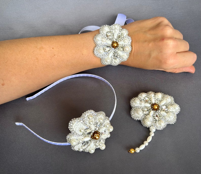Puede incluir: Un conjunto de tres broches de flores blancas de perlas y oro. Un broche est&aacute; unido a una diadema blanca, otro a una cinta blanca y el tercero a una cadena blanca.