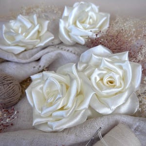 Può includere: Primo piano di rose in raso avorio, con petali sovrapposti, disposte su un tessuto neutro. I fiori fatti a mano sono di un bianco crema, con una lucentezza delicata. Sono visibili anche un gomitolo di lana e un ago.