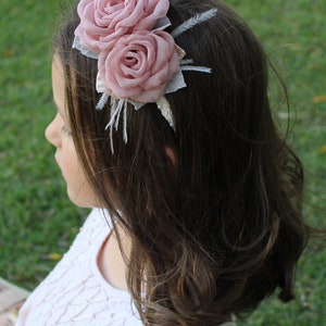 Op de afbeelding: Een roze bloemenhoofdband met twee stoffen rozen en witte veren. De hoofdband wordt gedragen door een persoon met lang bruin haar.