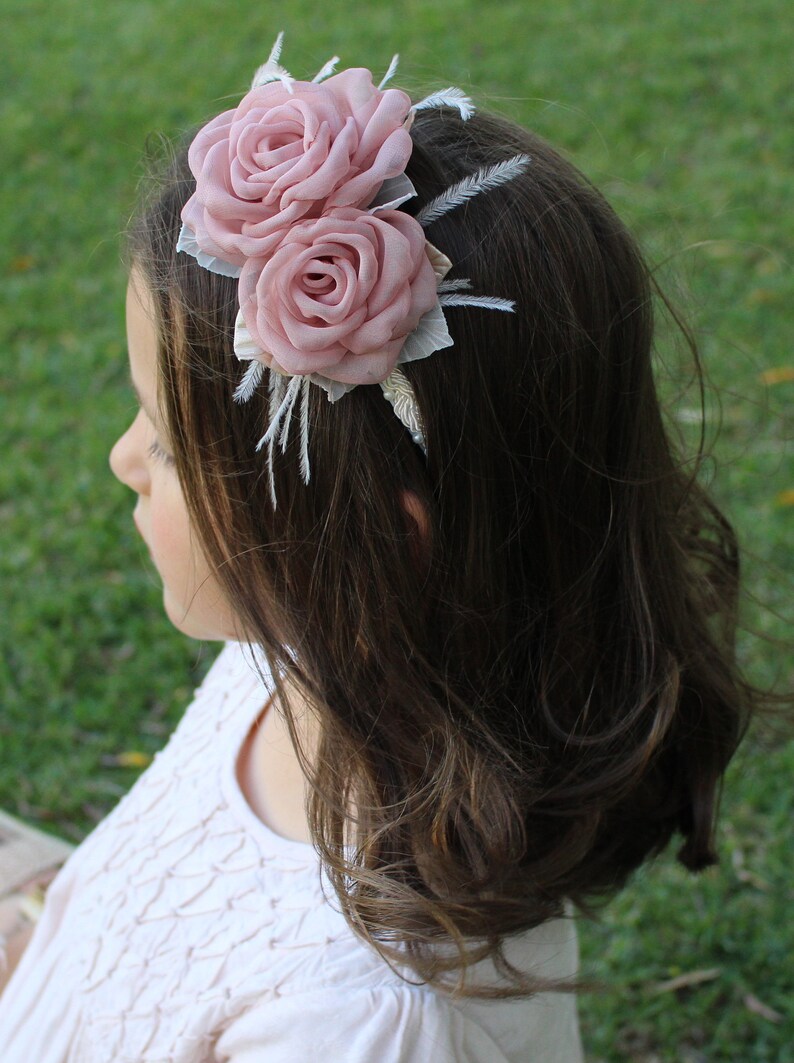 Op de afbeelding: Een roze bloemenhoofdband met twee stoffen rozen en witte veren. De hoofdband wordt gedragen door een persoon met lang bruin haar.
