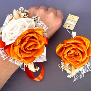 May include: A wrist corsage and boutonniere with orange and white satin roses and white baby's breath. The corsage is tied with an orange satin ribbon and has white lace trim.