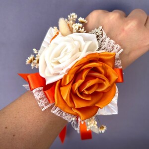 May include: A wrist corsage with a white satin rose and an orange satin rose, accented with white baby's breath and lace.