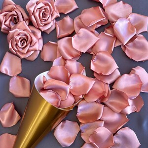 May include: A gold cone filled with rose petals, surrounded by loose rose petals on a gray surface. The petals are a soft, blush pink color and made of satin.
