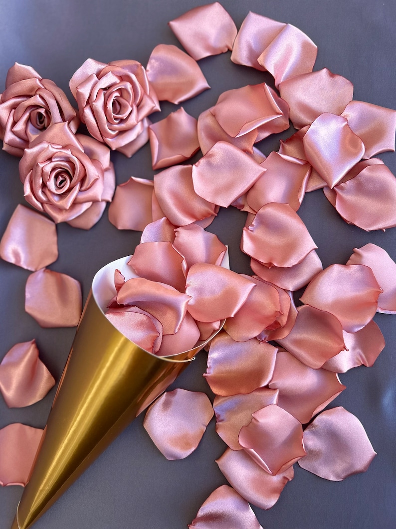 May include: A gold cone filled with rose petals, surrounded by loose rose petals on a gray surface. The petals are a soft, blush pink color and made of satin.