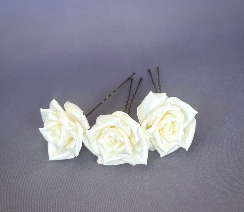 May include: Three white satin rose hair clips with silver bobby pins. The roses are arranged in a row, with the middle rose slightly larger than the other two.
