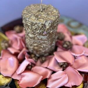 May include: A gold candle with a textured surface sits on a bed of pink satin rose petals. The candle is surrounded by small brown pine cones.