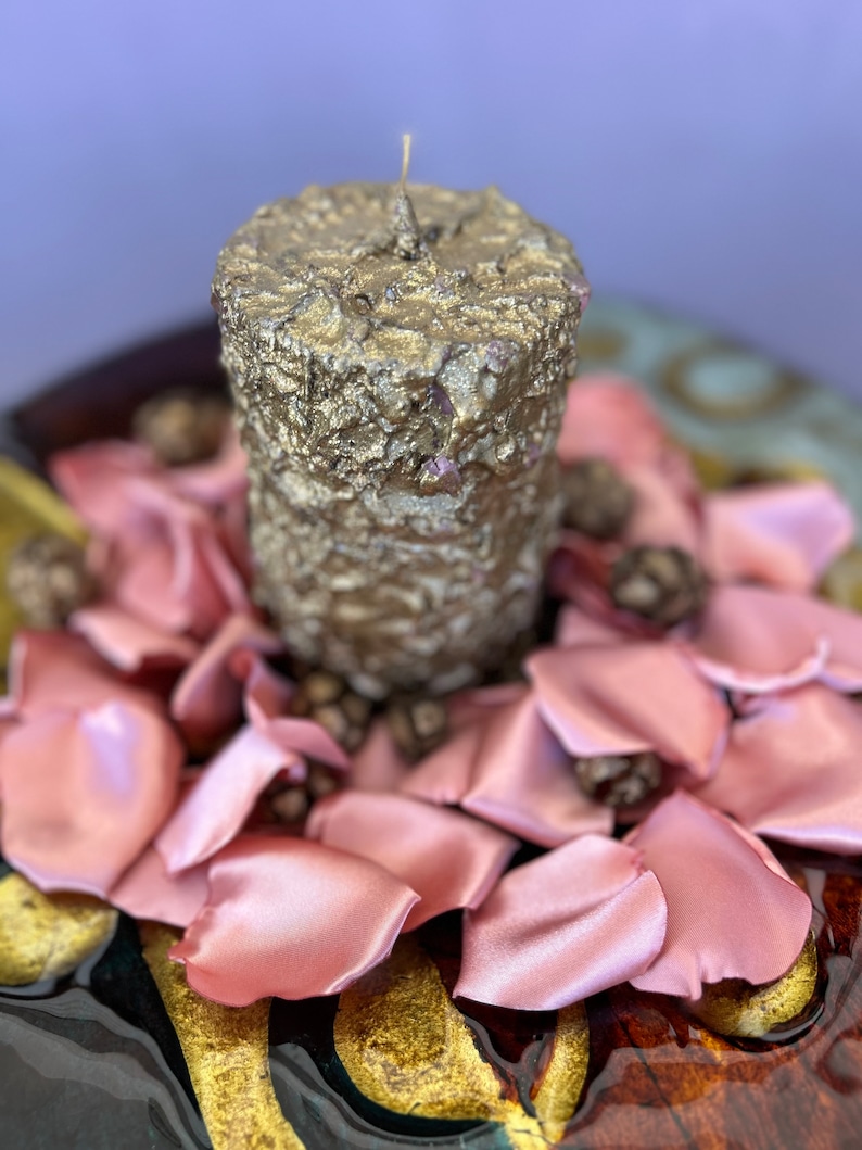 May include: A gold candle with a textured surface sits on a bed of pink satin rose petals. The candle is surrounded by small brown pine cones.