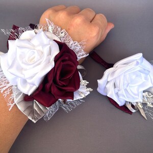 May include: A white and burgundy satin rose wrist corsage with lace and rhinestones. The corsage is tied with a burgundy ribbon.