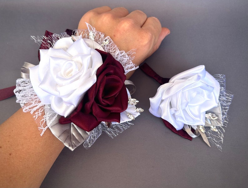 May include: A white and burgundy satin rose wrist corsage with lace and rhinestones. The corsage is tied with a burgundy ribbon.