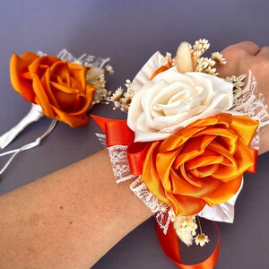 May include: A wrist corsage with orange and white satin roses, lace, and dried flowers. The corsage is tied with a long orange ribbon.