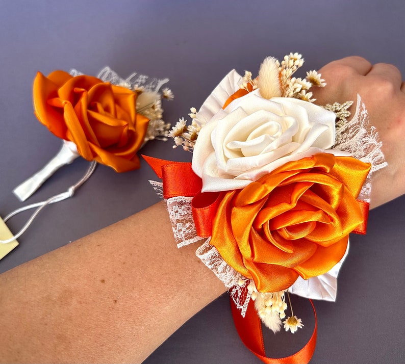 May include: A wrist corsage with orange and white satin roses, lace, and dried flowers. The corsage is tied with a long orange ribbon.
