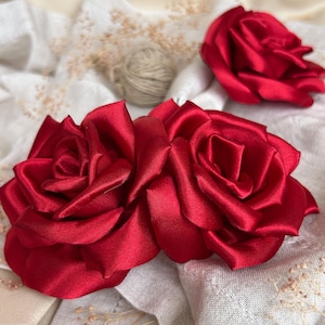 May include: Three deep red satin roses with layered petals arranged in a cluster. The flowers feature a smooth, reflective sheen and are positioned against a soft, off-white fabric background with small dried floral accents tucked between them.