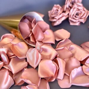 May include: A gold cone filled with rose petals, with three blush pink satin roses on a gray background.