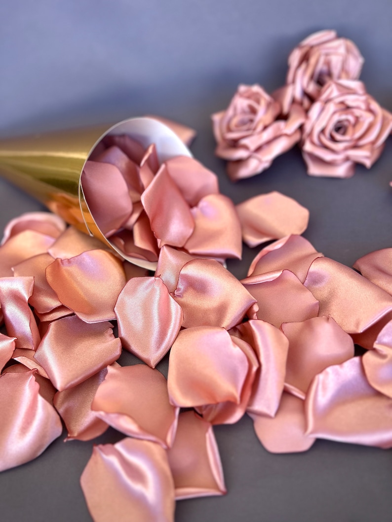 May include: A gold cone filled with rose petals, with three blush pink satin roses on a gray background.