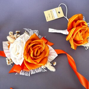 May include: A set of two orange and white silk flower boutonnieres with white lace and orange ribbon. The boutonnieres are perfect for a wedding or special occasion.