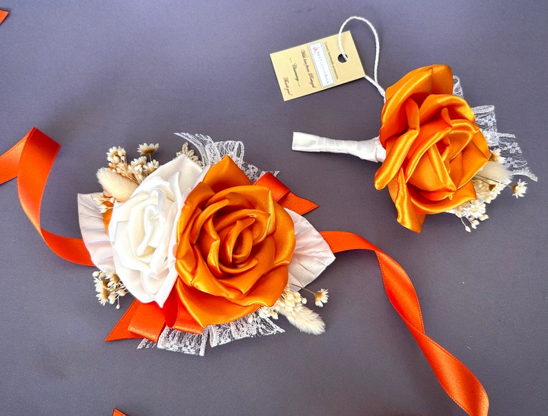 May include: A set of two orange and white silk flower boutonnieres with white lace and orange ribbon. The boutonnieres are perfect for a wedding or special occasion.