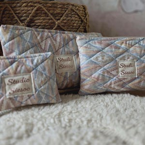 Op de afbeelding: Drie gewatteerde make-up tassen in verschillende maten, met een ruitpatroon in pastelkleuren blauw, roze en beige. Elke tas heeft een rechthoekig label met de tekst "Studio Siesoo". De tassen liggen op een witte, zachte ondergrond.
