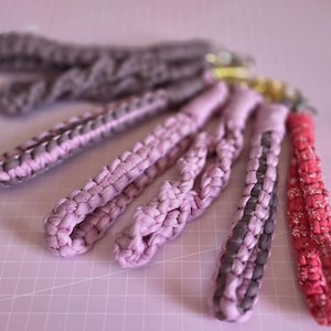 Op de afbeelding: Een verzameling van zes gevlochten stoffen polsband-sleutelhangers in diverse tinten roze, mauve en donkerbruin. Elke sleutelhanger heeft een geweven textiellus bevestigd aan een goudkleurige metalen draaisluiting met een getextureerd ontwerp.