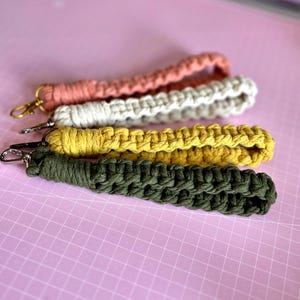 Op de afbeelding: Vier macramé polsband-sleutelhangers met geknoopt koord in terracotta, crème, mosterdgeel en olijfgroen. Elke polsband is afgewerkt met een goudkleurige metalen karabijnhaak bevestigd aan het uiteinde van de geweven stoffen lus.
