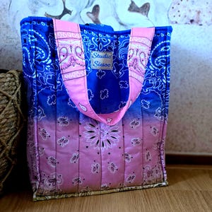 Op de afbeelding: Een gewatteerde draagtas met een bandana-print in een blauw naar roze verloop. De tas heeft twee roze hengsels en een rechthoekig label met de tekst Studio Siesoo op de voorzijde. De stof is voorzien van een herhalend paisley-patroon.