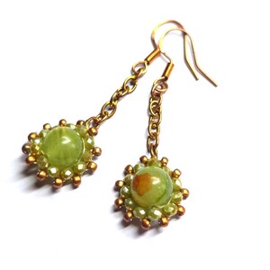 Può includere: Un paio di orecchini pendenti con una pietra verde centrale, circondata da perline più piccole verde chiaro e dorate. Gli orecchini sono collegati da una catena dorata e hanno ganci per le orecchie dorati. Il design è floreale.