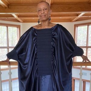 May include: A navy blue, flowing dress with a pleated bodice and wide sleeves. The dress is made of a shiny, satin-like fabric. The person is standing on a wooden floor with a white railing.