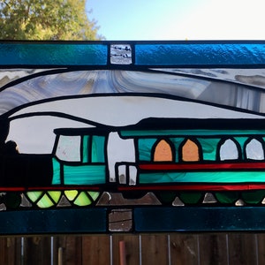 May include: A stained glass window depicting a green and black train with white smoke. The train has a red stripe and multiple windows. The background is a light blue.