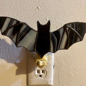 May include: A black and white stained glass bat nightlight plugged into an electrical outlet. The bat has a black body and wings with white stripes.