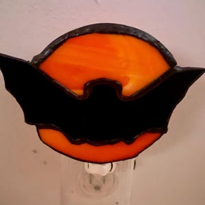 May include: A stained glass night light featuring a black bat silhouette against an orange background. The bat is centered, with its wings spread, and the orange glass is visible through the wings. The night light is plugged into a white outlet.